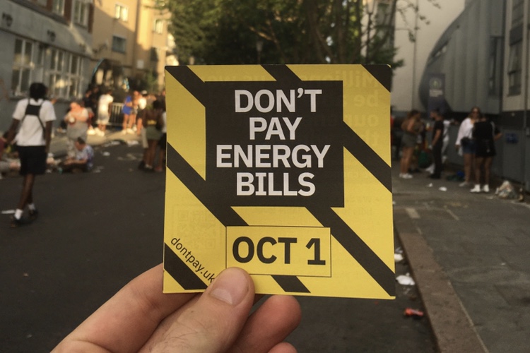 A flyer for the Don't Pay campaign, which reads "Don't Pay Energy Bills - October 1st' against a striped black and yellow background. The flyer is held in front of a crowd at Notting Hill Carnival.